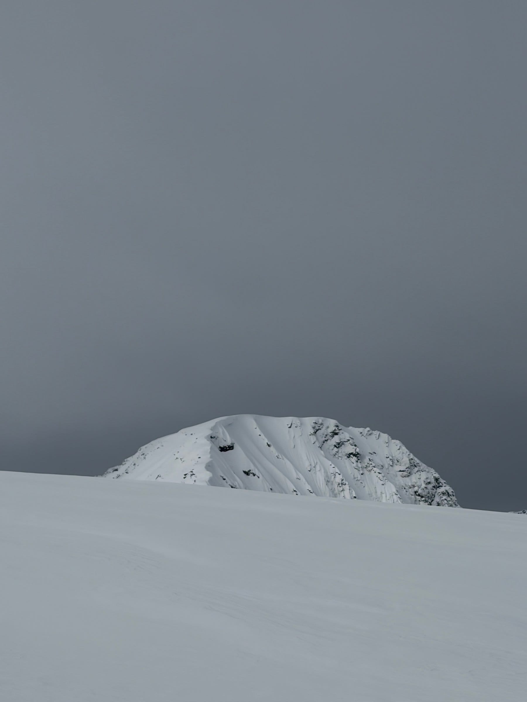 En snødekt bergtopp stiger opp fra et hvitt snølandskap under en tung, grå overskyet himmel.