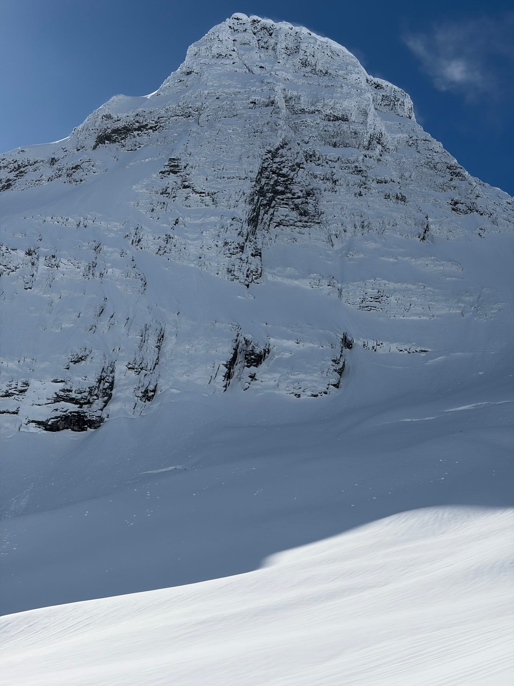 En bratt snødekt fjelltopp med dramatiske bergvegger reiser seg mot en klar blå himmel, med et glatt vindpakket snøfelt i forgrunnen.