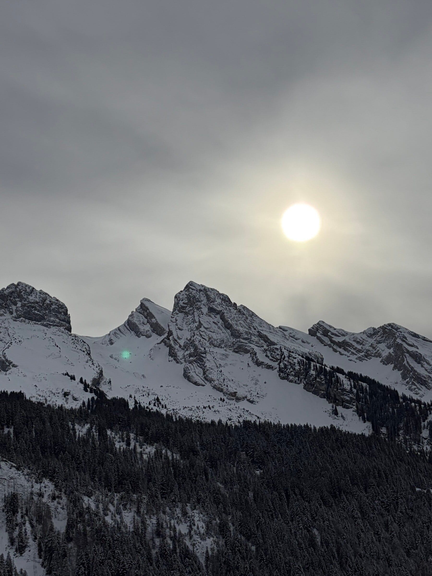 Solen lyser bak en steinete fjelltopp med snø og trær